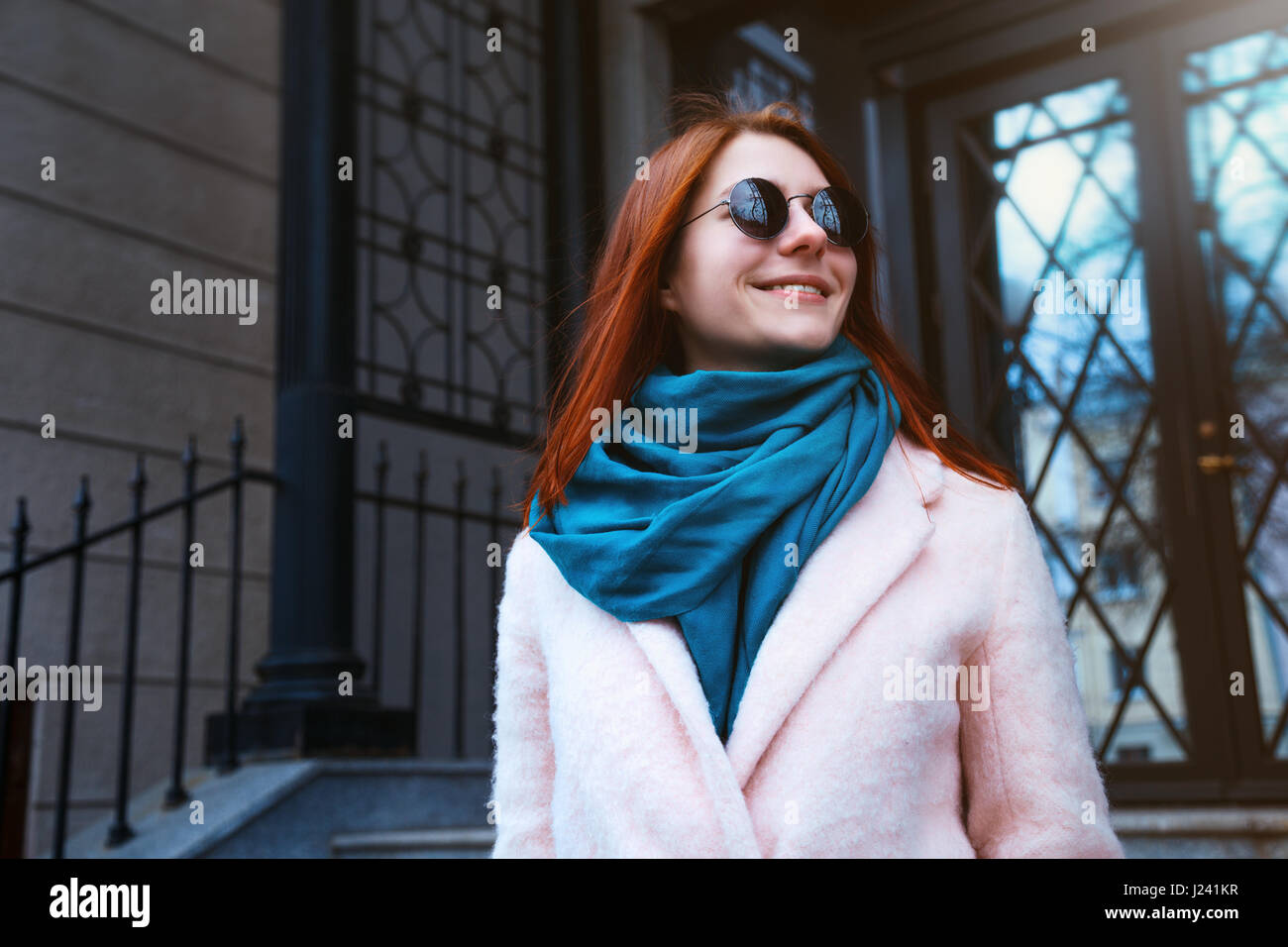 Dai Capelli rossi ragazza bella sta camminando per strada in un cappotto rosa e blu sciarpa, con occhiali da sole. Foto Stock