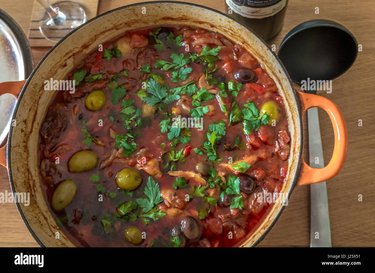 Grandi ghisa vaso riempito con stufato spagnolo, olive, pollo, patate, il coriandolo, il prezzemolo, pomodori secchi, servita su tavola in legno di quercia con siviera Foto Stock