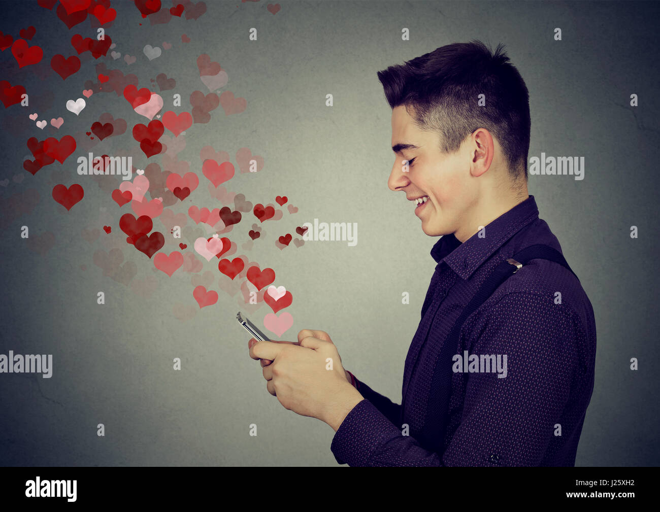 Profilo laterale uomo felice amore invio messaggio di testo sms sul cellulare con cuori rossi volare lontano dallo schermo isolato sul muro grigio Sfondo. Positiv Foto Stock