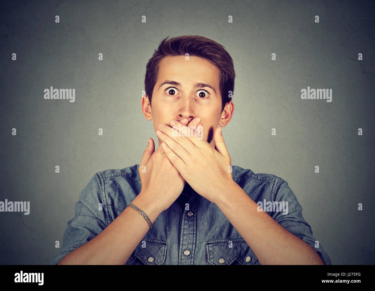 Spaventata giovane uomo che copre la bocca con le mani isolato sul muro grigio Sfondo. Emozioni umane espressione faccia Foto Stock
