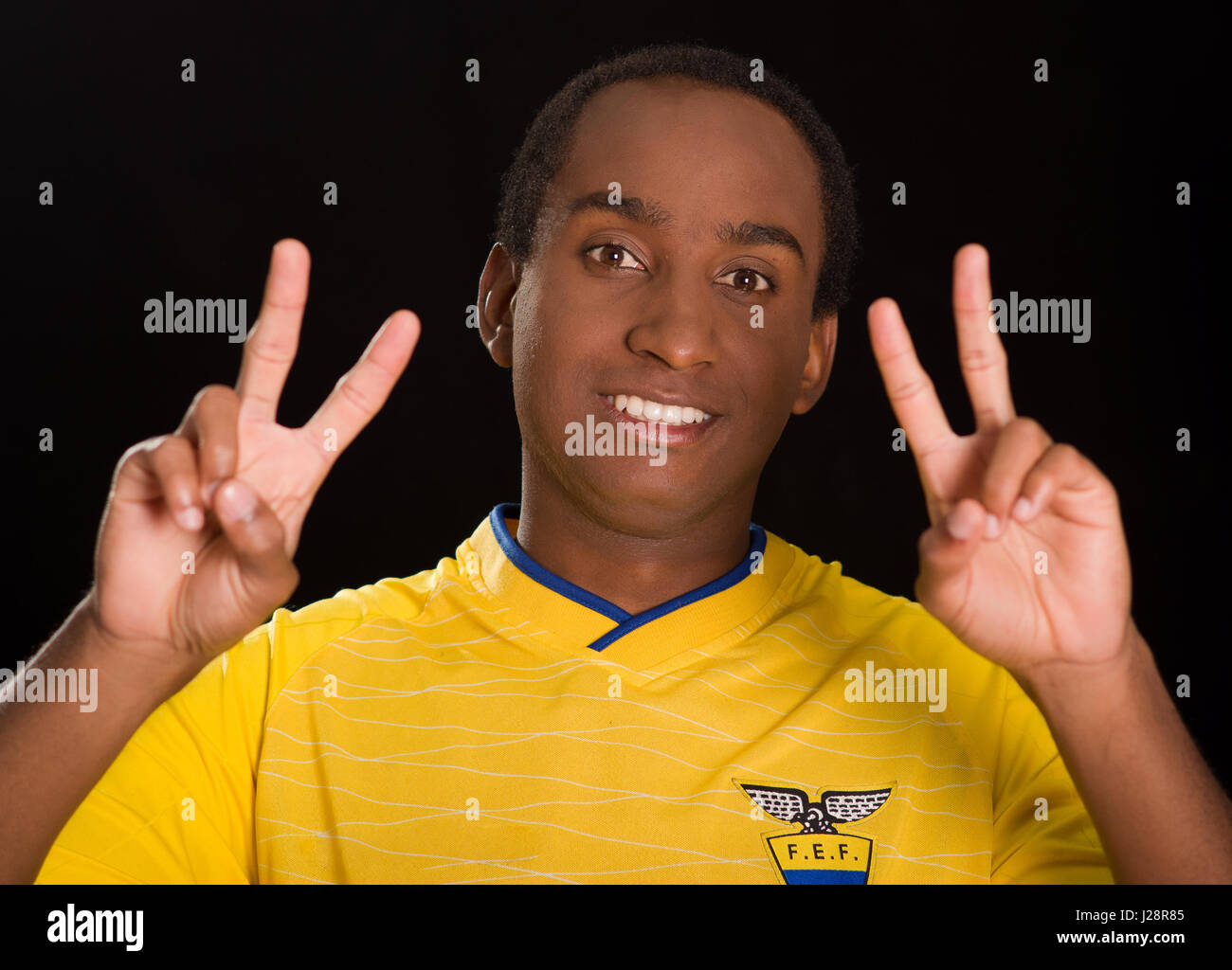 Quito, Ecuador - 25 Maggio 2016: Headshot dark scuoiati maschio giallo da indossare ecuadoriano football shirt davanti a sfondo nero, tenendo su entrambe le mani facendo segni di pace sorridente Foto Stock