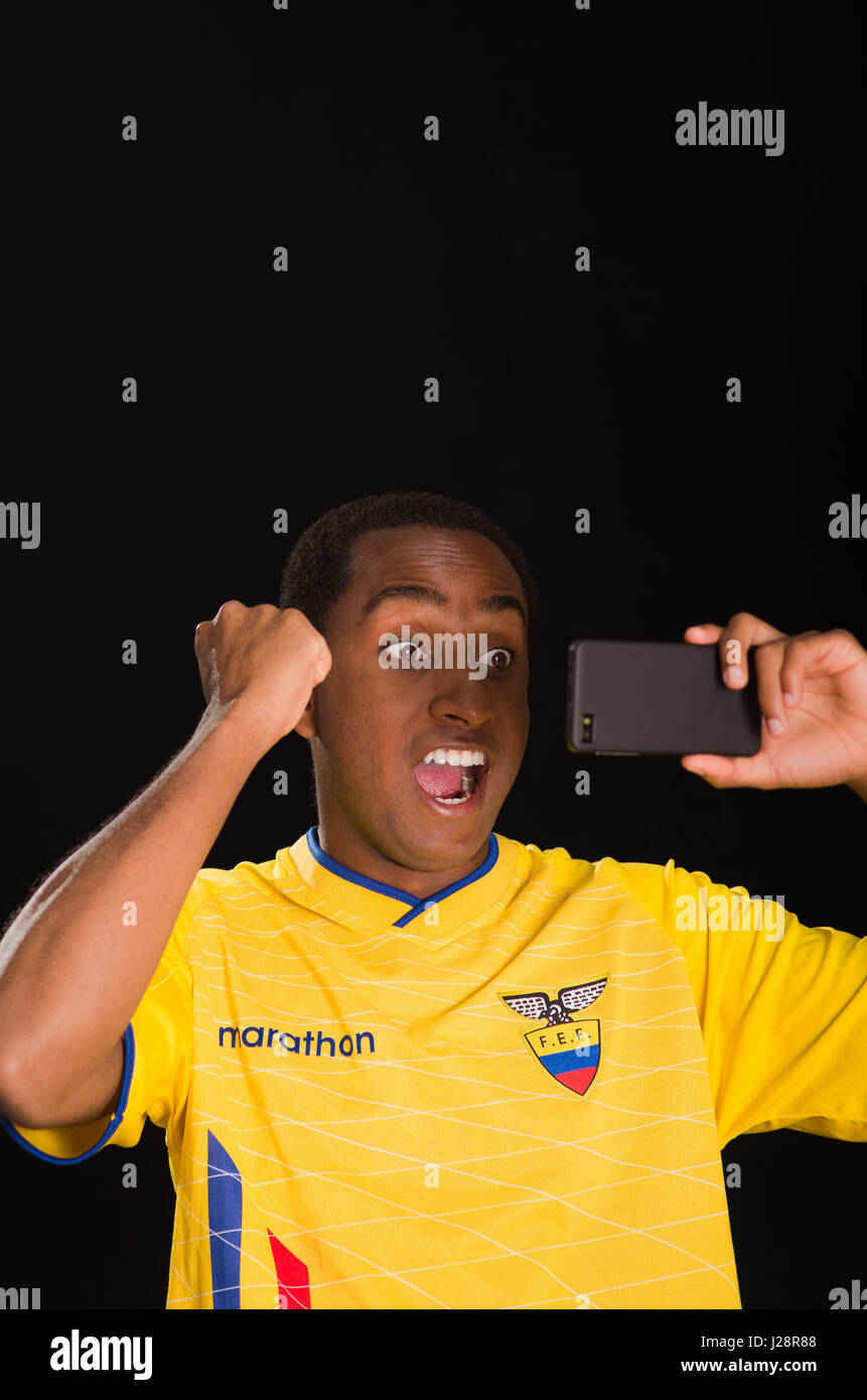 Quito, Ecuador - 25 Maggio 2016: Headshot dark scuoiate indossando maschio giallo ecuadoriana football shirt davanti a sfondo nero, tenendo in mano il telefono cellulare guardando lo schermo e il tifo con entusiasmo Foto Stock