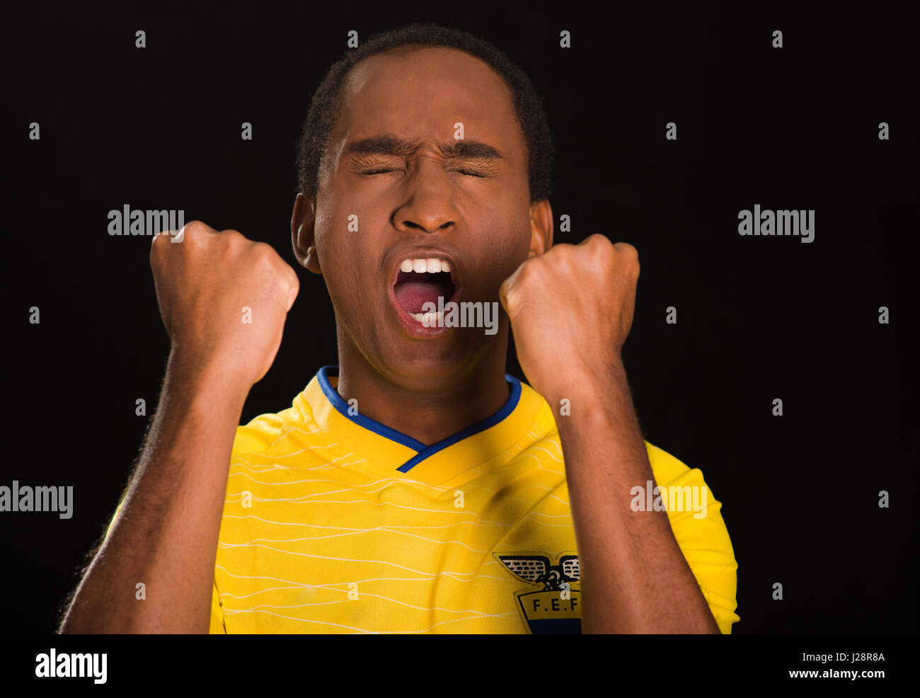 Quito, Ecuador - 25 Maggio 2016: Headshot dark scuoiati maschio giallo da indossare ecuadoriano football shirt davanti a sfondo nero, gli occhi chiusi e la bocca aperta il tifo con le braccia sollevate Foto Stock