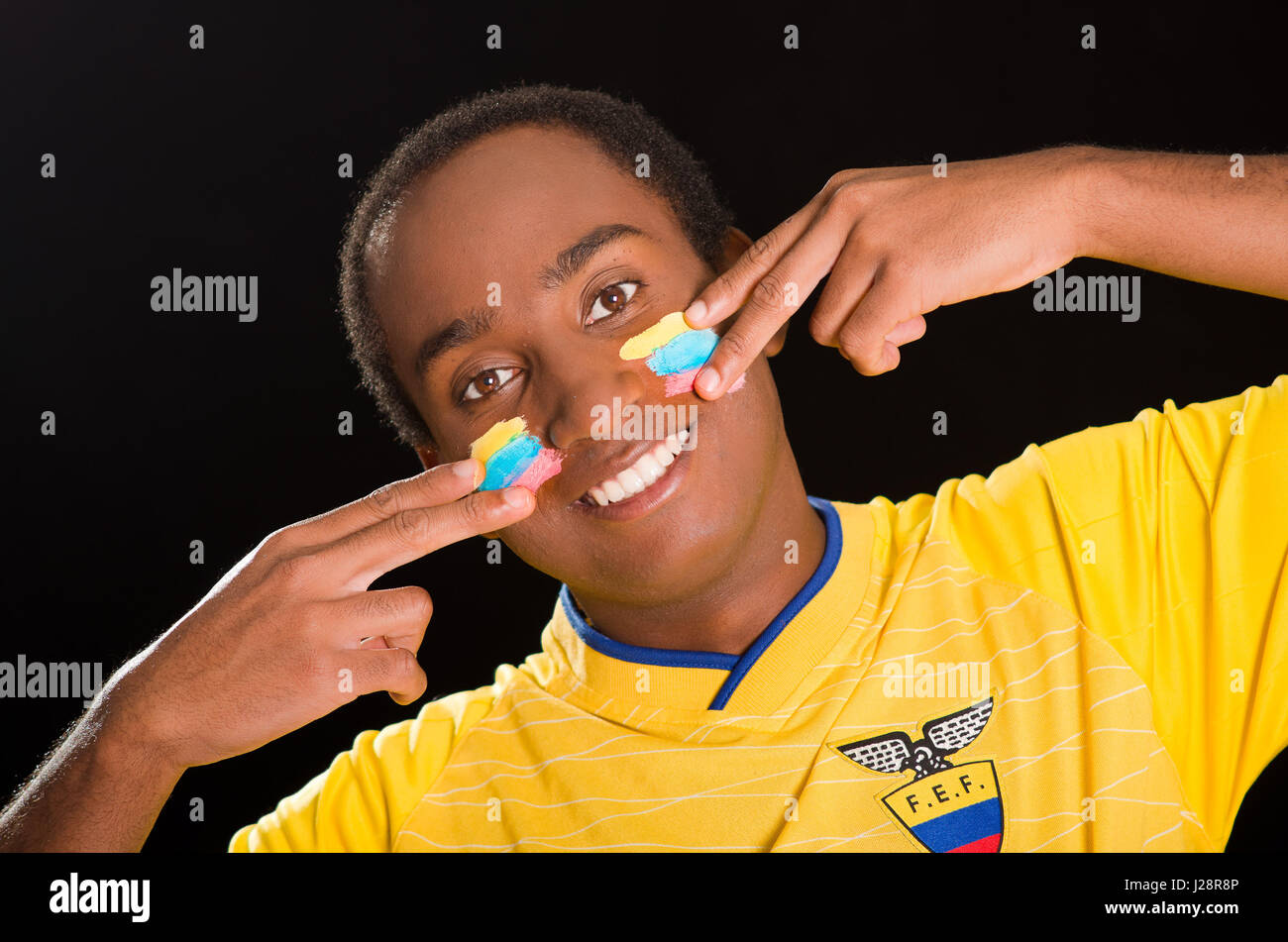 Quito, Ecuador - 25 Maggio 2016: Headshot dark scuoiate indossando maschio giallo ecuadoriana football shirt davanti a sfondo nero, usando le dita applicando facepaint guance Foto Stock