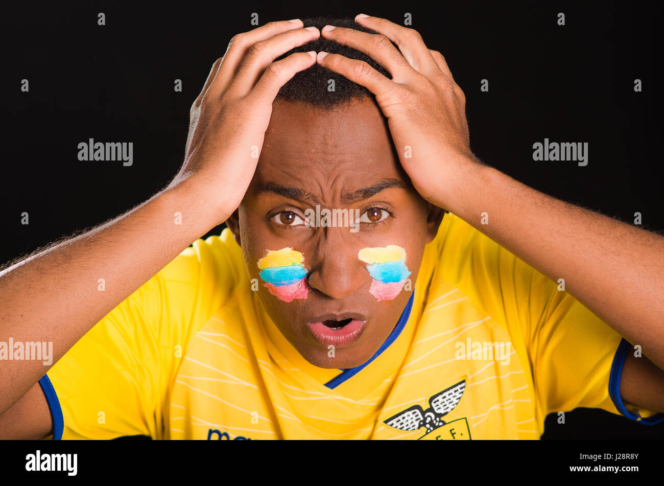 Quito, Ecuador - 25 Maggio 2016: Headshot dark scuoiati maschio giallo da indossare ecuadoriano football shirt davanti a sfondo nero, bandiera vernice per il viso, rivolto verso la fotocamera e il tifo con entusiasmo Foto Stock