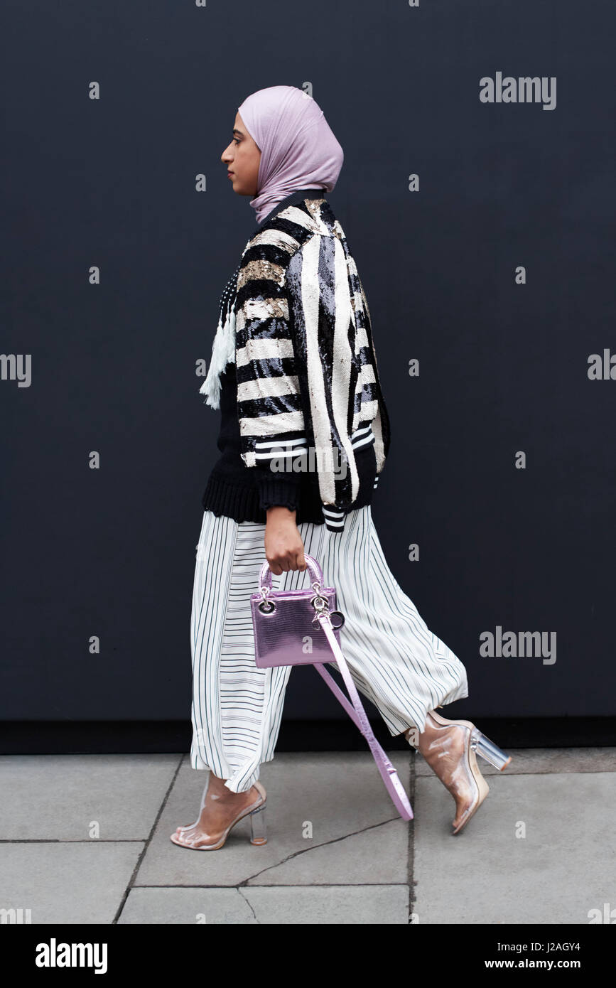 Londra - febbraio, 2017: a lunghezza piena vista della donna che cammina in una strada che indossa un hijab con bianco e nero a strisce top, ampi pantaloni e trasparente e di alta scarpe tacco, tenendo un piccolo metallico lilla Dior borsetta, London Fashion Week, giorno 5. Foto Stock