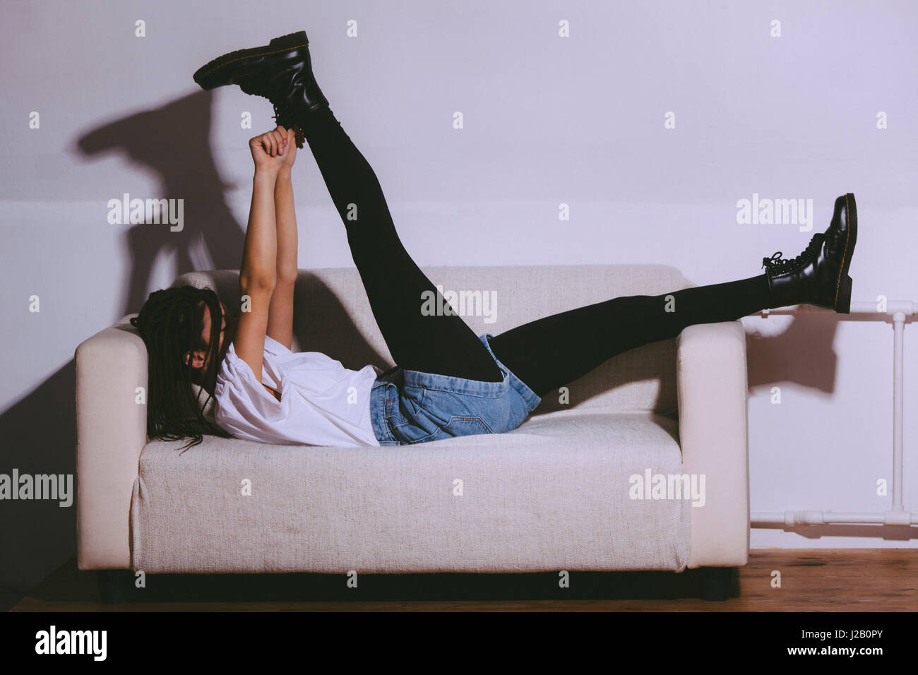 Vista laterale di giocosa donna sdraiata sul divano mentre il cassone di contenimento contro il muro bianco Foto Stock