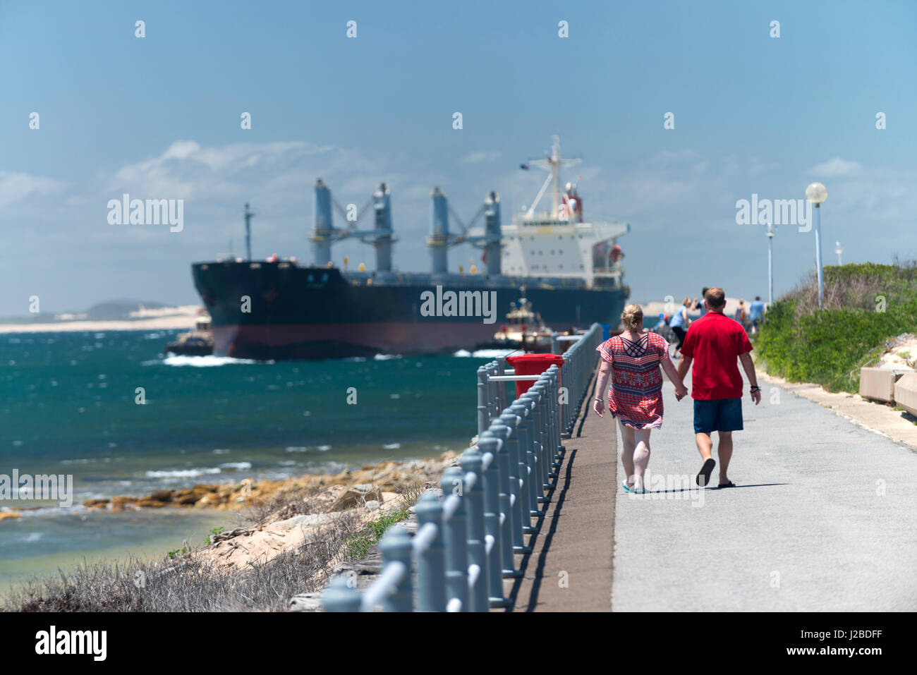 Una grande nave di carbone passa Nobbys Testa e walkers come lascia l'Oceano Pacifico ed entra nel porto di Newcastle e il Cacciatore estuario del fiume, Australia. Foto Stock