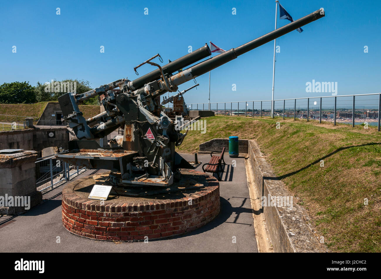 Un leggermente arrugginite da 3,7 pollici anti-aerei pistola punti su una banca erbosa verso gli edifici e la spiaggia di una località balneare bagnata dal sole estivo Foto Stock