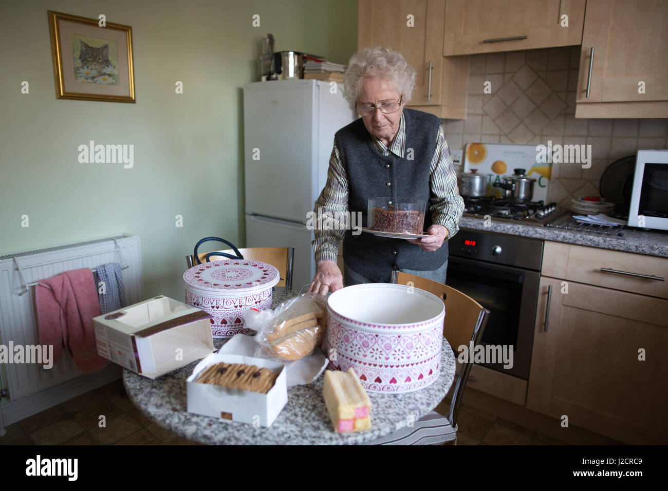 Signora anziana preparare un tè inglese con vari tipi di torte sul suo tavolo da cucina, England, Regno Unito Foto Stock