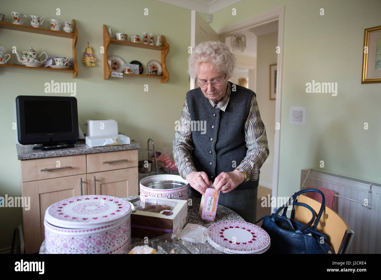 Signora anziana preparare un tè inglese con vari tipi di torte sul suo tavolo da cucina, England, Regno Unito Foto Stock