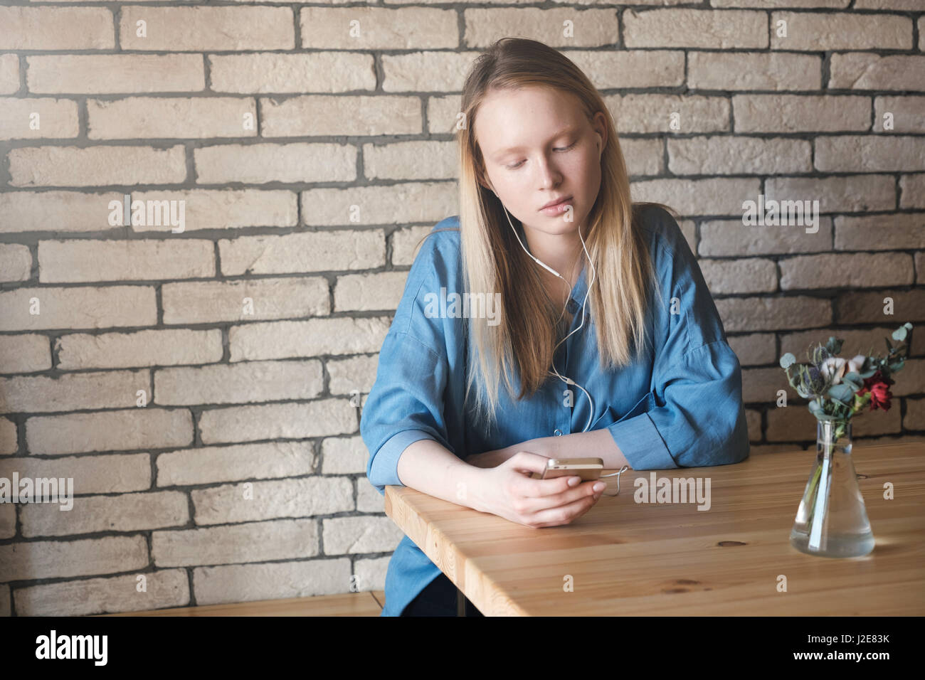 Triste ragazza bionda in blu camicetta seduti ad un tavolo in un cafe, ascolto di musica con cuffie e guardando al telefono. Foto Stock