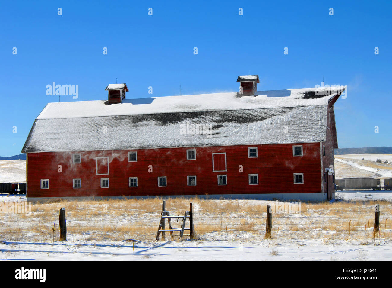 Il carbone treno viaggia dietro il vecchio fienile in legno in Colorado. Il vento soffia neve dal tetto del granaio rosso. Foto Stock