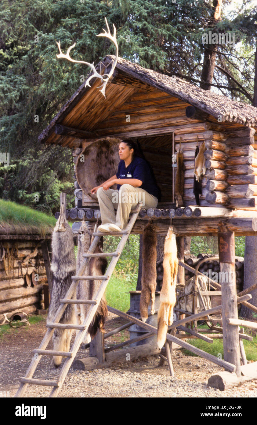 Athabaskan donna siede sulla piattaforma di un registro tradizionalmente costruite nella conservazione di alimenti shelter, chiamata cache, situato all'interno di un caccia e pesca camp. Villaggio Chena, Alaska Foto Stock