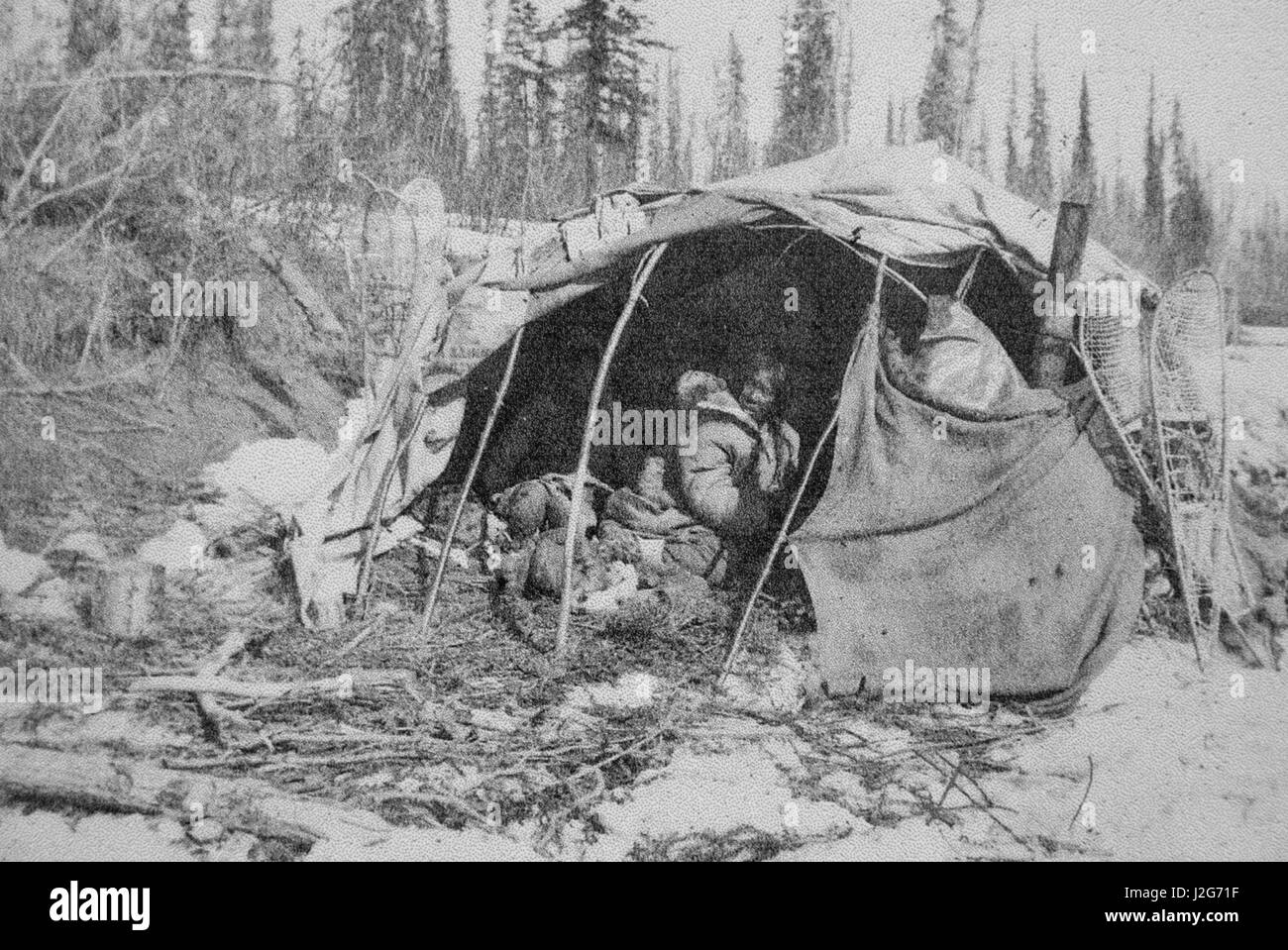 Storica fotografia in bianco e nero di un Athabaskan dimora di piegate alberello telaio e ricoperto con tela e coperte. Coppia di racchette da neve si appoggia contro la struttura. Alaska Foto Stock