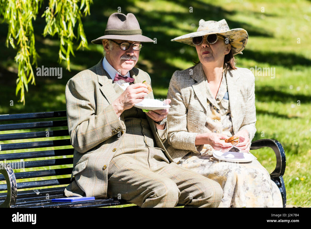 Saluto alla 40s evento. Una coppia senior su una panchina nel parco pausa loro panini di posa per una foto. Entrambi vestiti in circa 1940 costume. Foto Stock