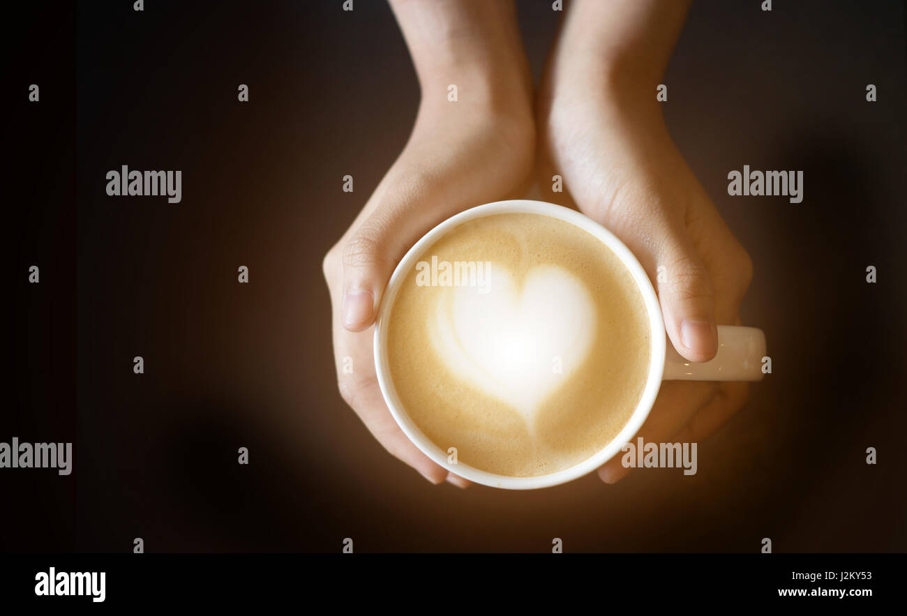 Latte Art come forma di cuore in tazza di caffè con tenere in mano la tazza per il caldo in uno sfondo scuro Foto Stock