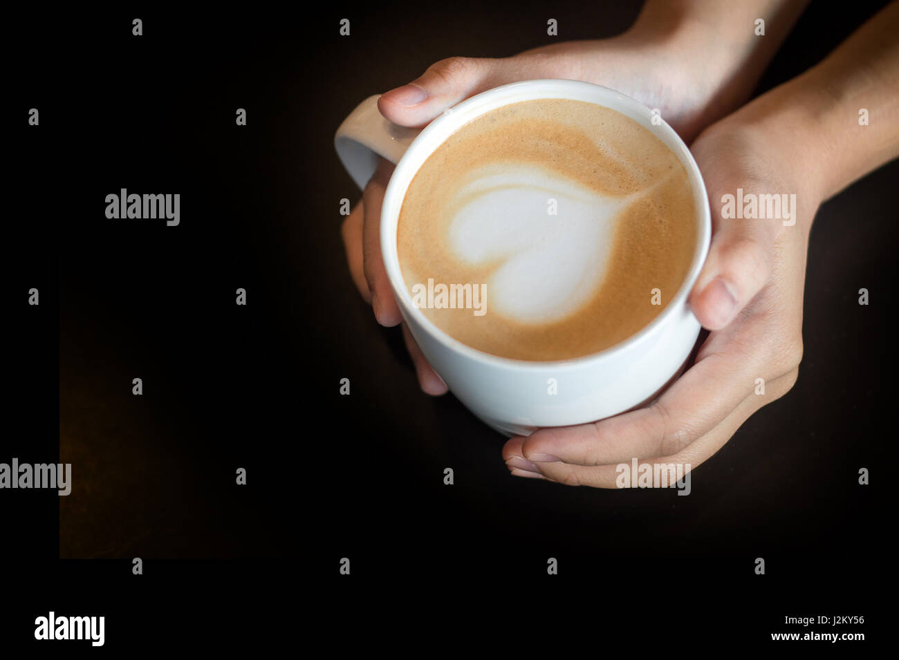 Latte Art come forma di cuore in tazza di caffè con tenere in mano la tazza per il caldo in uno sfondo scuro Foto Stock