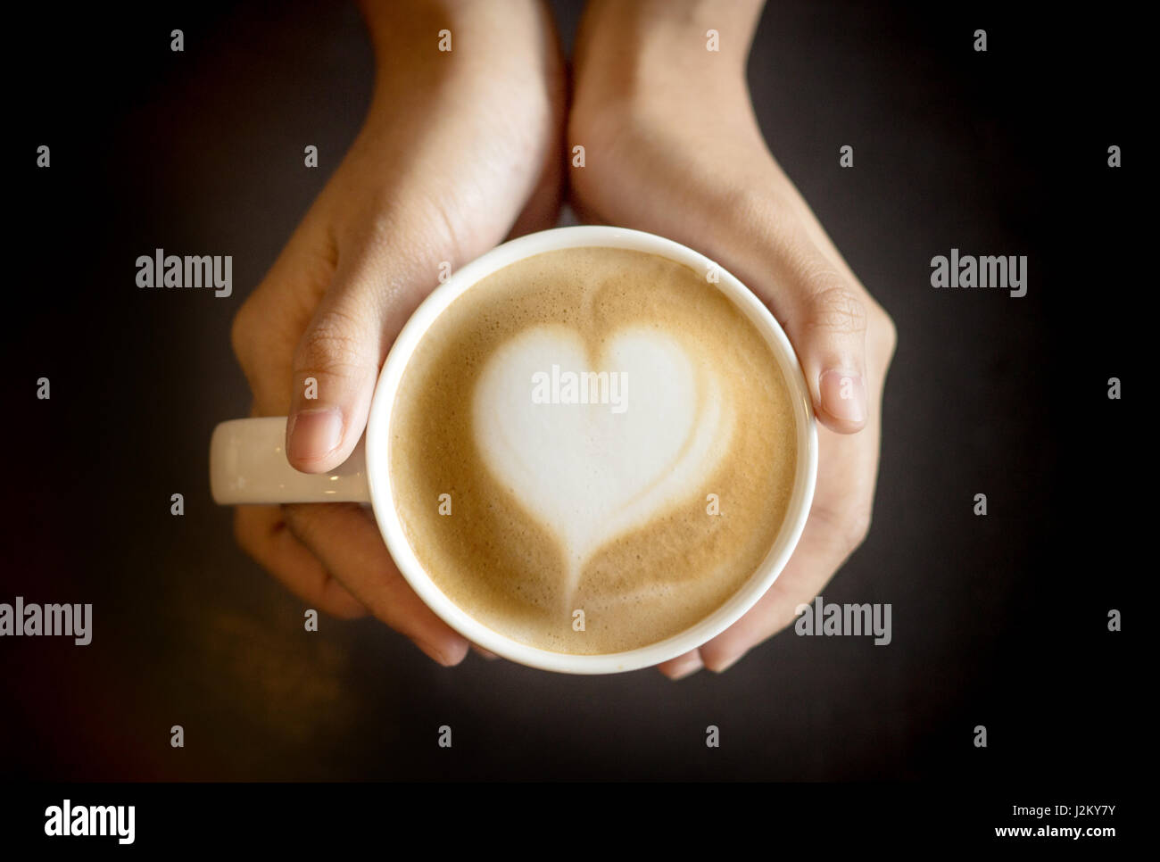 Latte Art come forma di cuore in tazza di caffè con tenere in mano la tazza per il caldo in uno sfondo scuro Foto Stock