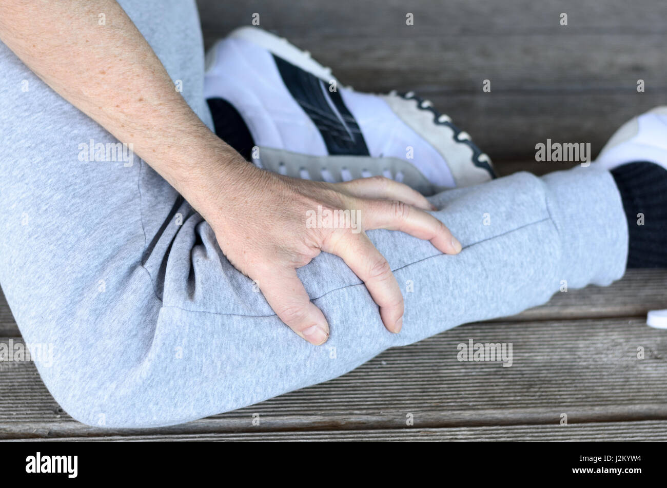 Sportivo con uno spasmo muscolare o crampi nel polpaccio stringendo la sua gamba inferiore con la sua mano in una vista ravvicinata su una piattaforma di legno Foto Stock