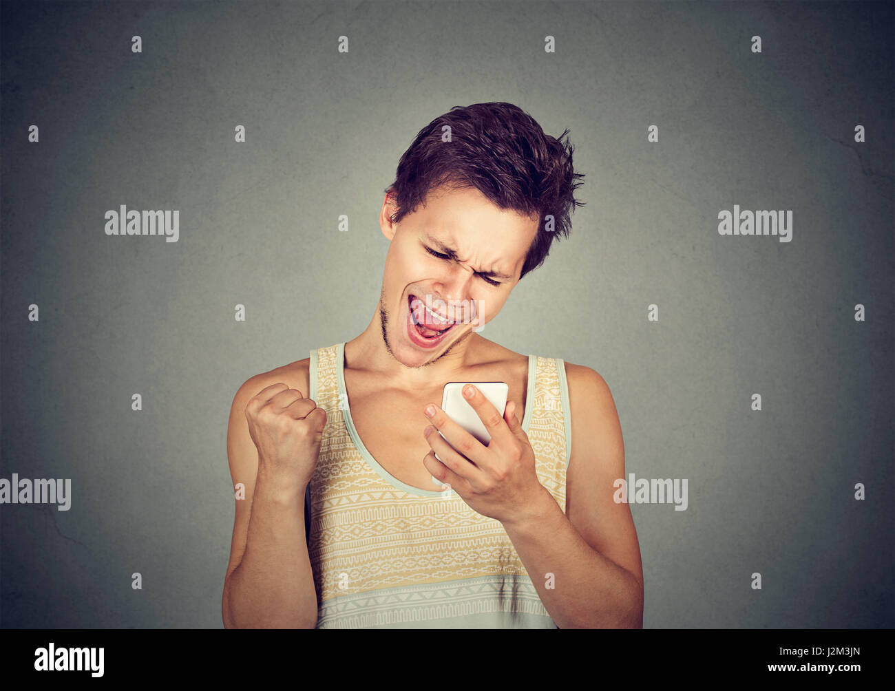 Restare connessi. Felice giovane guardando al suo telefono mobile e sorridente pugno di pompaggio celebra il successo isolato su sfondo grigio. Laughi eccitato Foto Stock