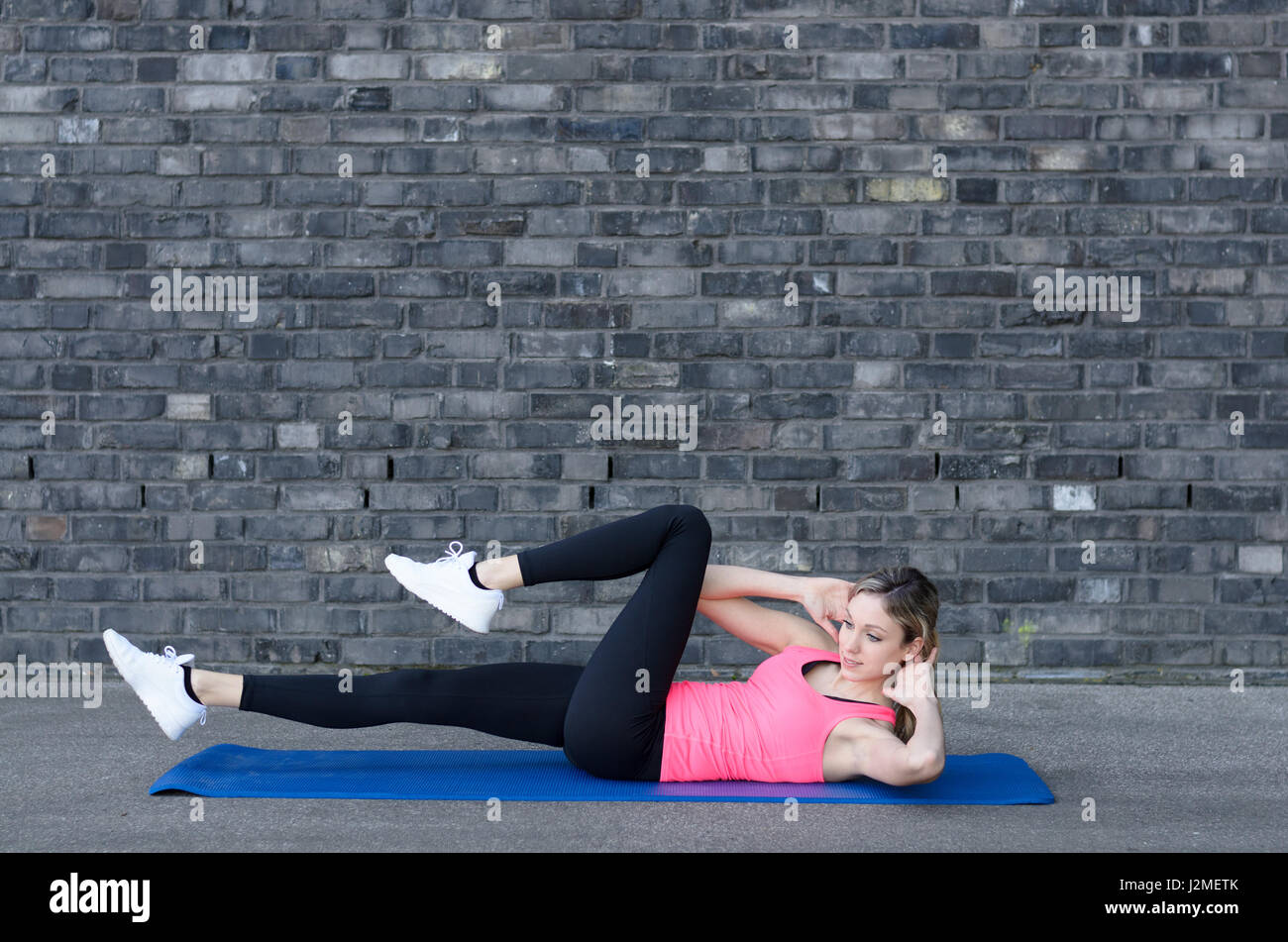 Montare donna sorridente guardando sopra la spalla mentre facendo esercizi addominali con alternanza di estensioni di gamba grigio vicino a un muro di mattoni Foto Stock