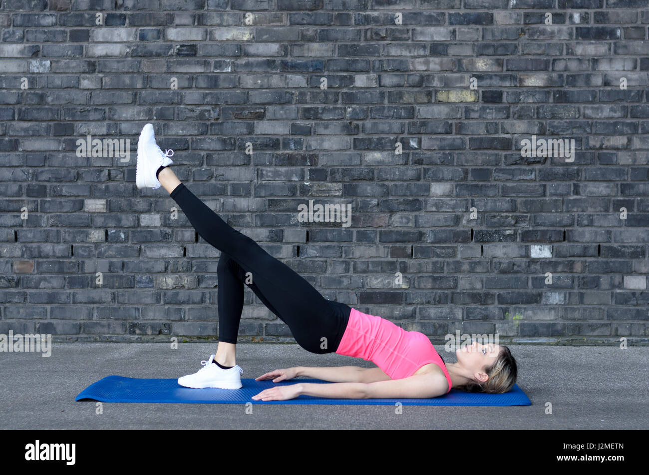 Montare donna sorridente guardando sopra la spalla mentre facendo esercizi addominali con alternanza di estensioni di gamba grigio vicino a un muro di mattoni Foto Stock