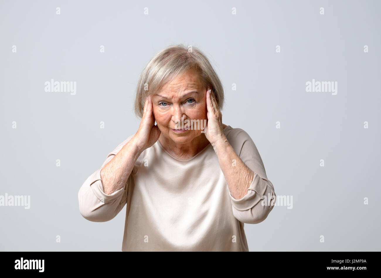 Senior grey-haired donna toccando i suoi templi con ambedue le mani alla sua testa, purtroppo guardando la telecamera come se soffrono di mal di testa, in piedi contro Foto Stock