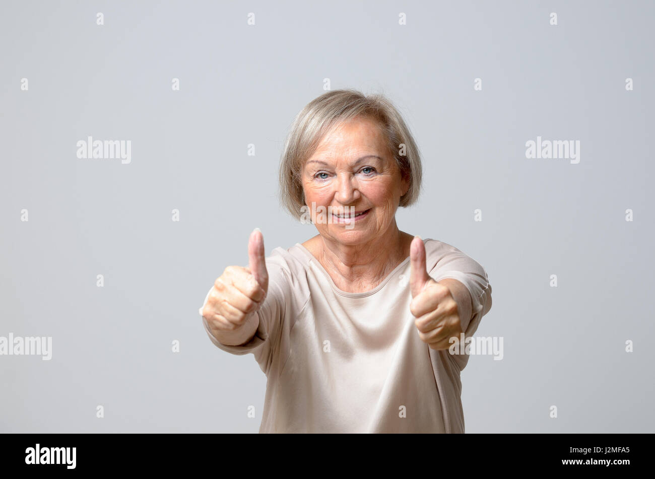 Senior donna nella sua 70s mostra pollice su gesti con entrambe le mani, guardando la fotocamera e sorridente, in piedi contro la pianura sfondo grigio Foto Stock