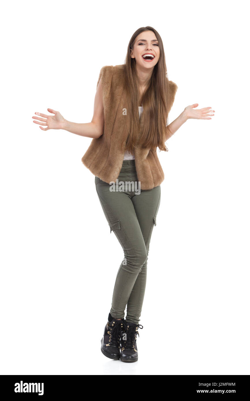 Bella giovane donna in pelliccia marrone gilet, i pantaloni khaki e stivali neri in piedi con le braccia tese, ridendo e guardando la fotocamera. A piena lunghezza Foto Stock
