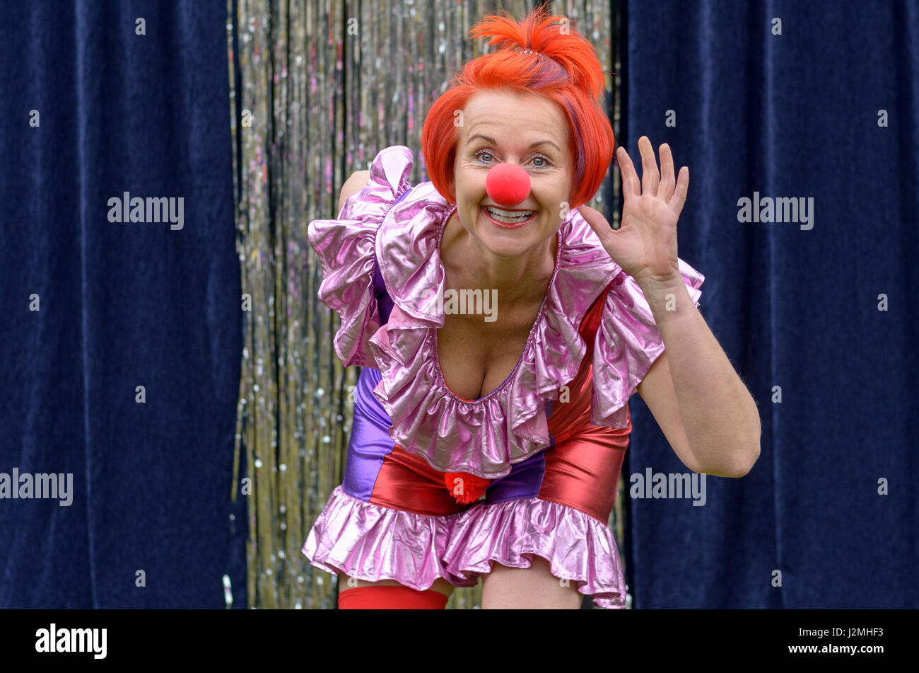 Carino divertente redhead attore in un rosso clown naso e vivacemente colorato costume flessione in avanti sventolando la fotocamera con un vivace grin Foto Stock