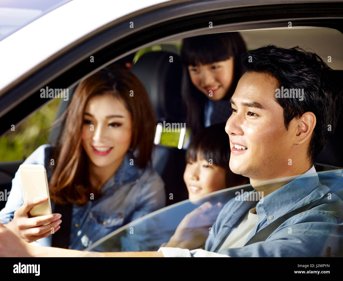 Felice famiglia asiatica con due bambini che viaggiano in auto, madre usando il cellulare mentre padre guida, concentrarsi sul padre. Foto Stock