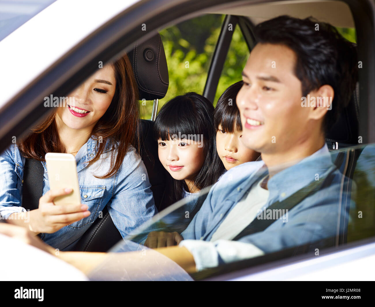Felice famiglia asiatica con due bambini che viaggiano in auto, madre usando il cellulare mentre padre guida, concentrarsi su la bambina. Foto Stock
