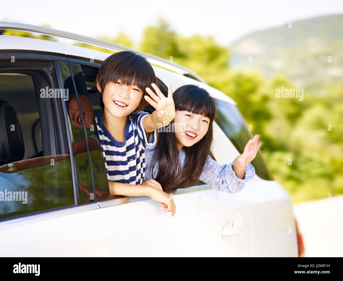 Felici i bambini asiatici teste di incollaggio al di fuori del finestrino posteriore di una vettura e agitando le mani. Foto Stock