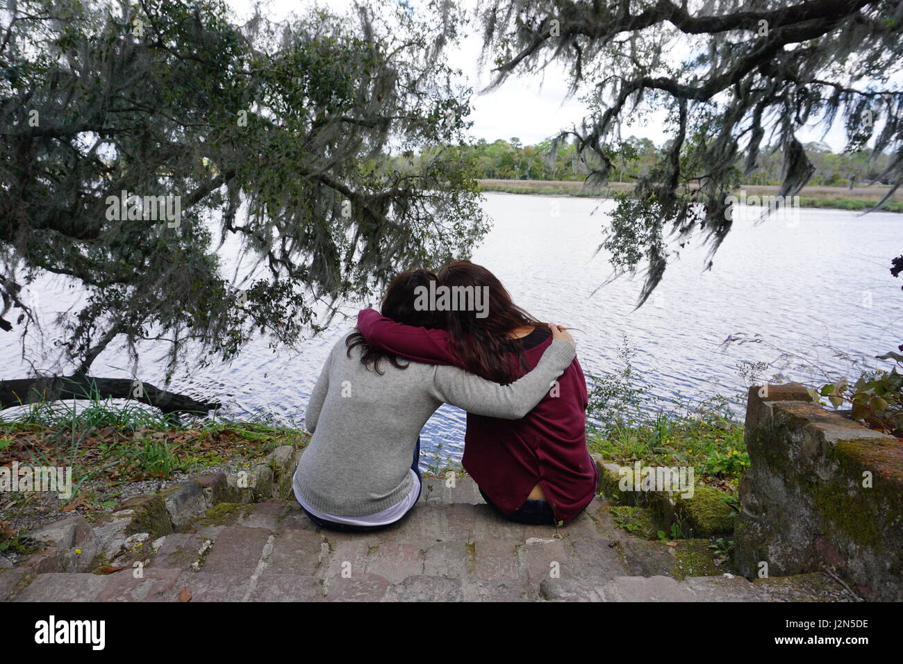 Due giovani donne sono seduti insieme rivolto indietro in un abbraccio fraterno come amici o sorelle guardando una laguna durante una mezza giornata di cammino. Foto Stock