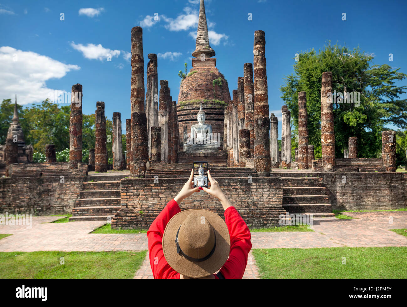 Tourist donna in costume rosso di scattare una foto del Buddha antico con il suo smartphone in Wat Sa Si di Sukhothai Historical Park, Thailandia Foto Stock