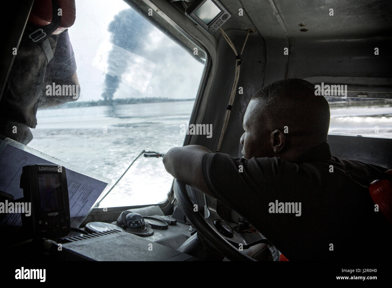 Il capitano di un piccolo cannoniere naviga verso il basso il bello fiume come flutti di fumo da una raffineria illegale sul fuoco, fiumi stato, Nigeria Foto Stock