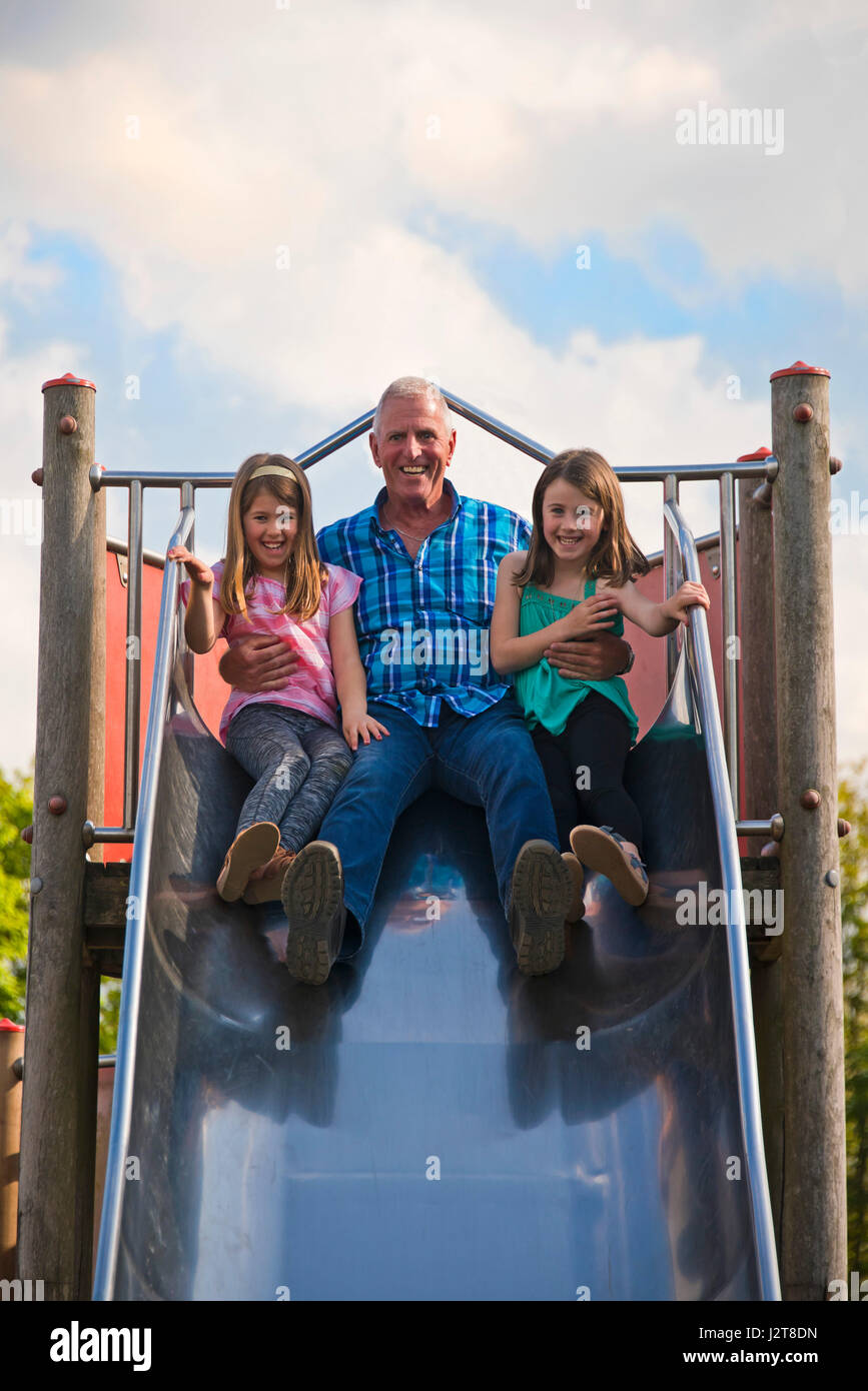 Ritratto verticale di un nonno lo scorrimento verso il basso una diapositiva con i suoi nipoti in un parco giochi. Foto Stock