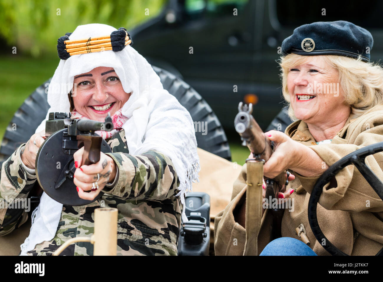 Inghilterra, sandwich. Saluto alla 40s evento. Due ridere donne mature seduta nella guerra mondiale due jeep, uno in possesso di una pistola di tommy e averlo al viewer.Si indossa un berretto, gli altri paesi arabi, copricapo. Foto Stock
