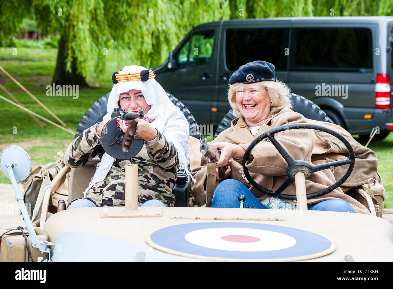 Inghilterra, sandwich. Saluto alla 40s evento. Due ridere donne mature seduta nella guerra mondiale due jeep, uno in possesso di una pistola di tommy e averlo al viewer.Si indossa un berretto, gli altri paesi arabi, copricapo. Foto Stock