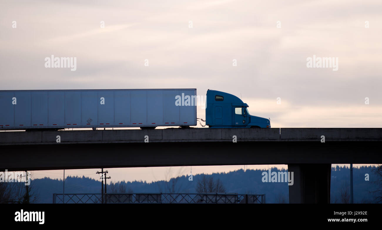 Silhouette di moderno semi-carrello con una traversina alta cabina e un lungo il rimorchio su un ponte di cemento costruito sull'entrata dell'autostrada di sera Foto Stock