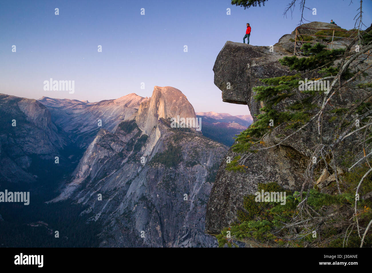 Un intrepido escursionista è in piedi su una roccia a strapiombo godendo della vista verso il famoso Half Dome presso il Glacier Point si affacciano in Twilight, Yosemite NP, STATI UNITI D'AMERICA Foto Stock