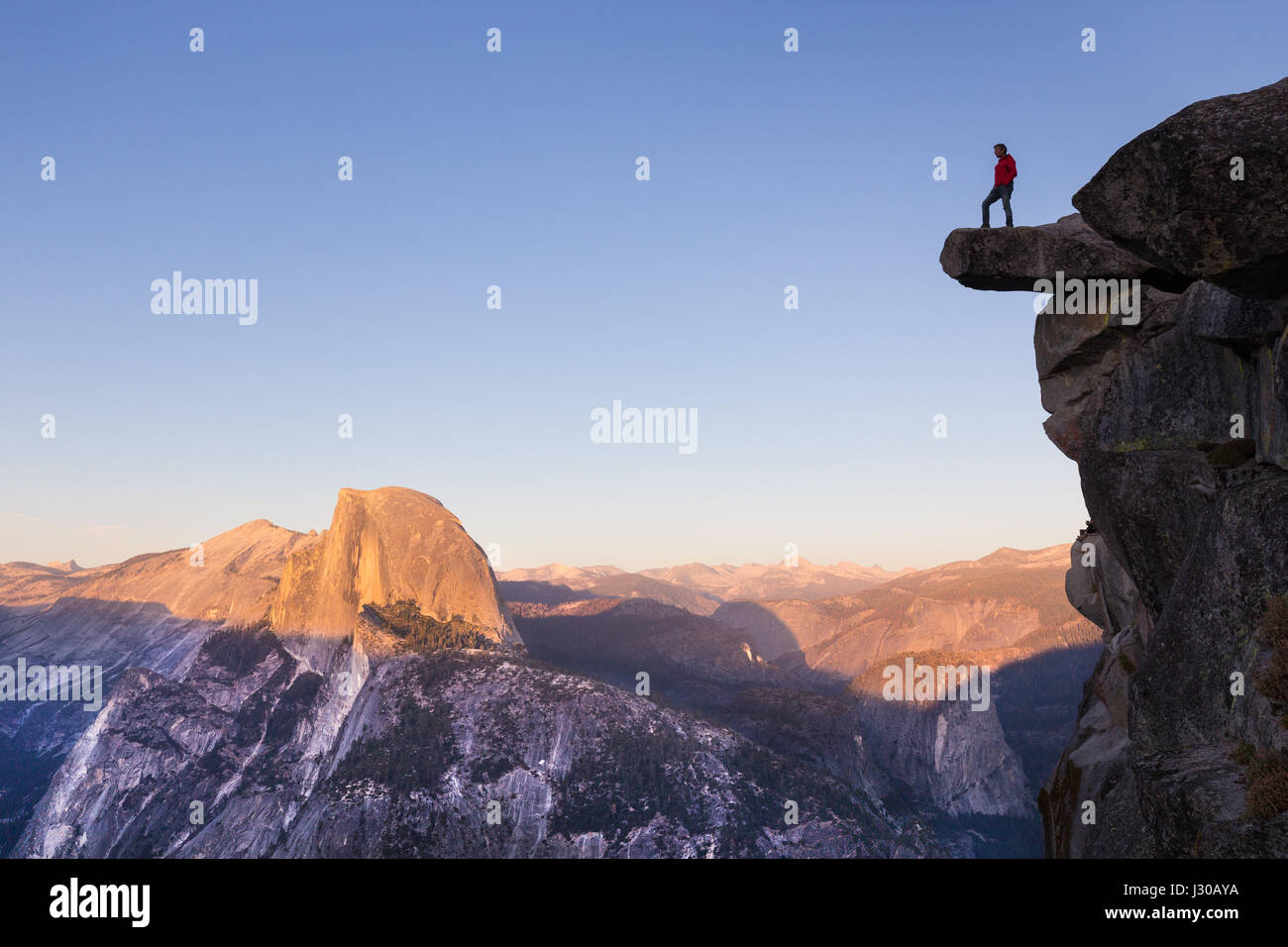 Un intrepido escursionista è in piedi su una roccia a strapiombo godendo della vista verso il famoso Half Dome presso il Glacier Point si affacciano nel bellissimo tramonto t Foto Stock