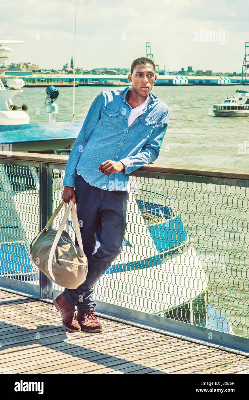 In viaggio. Indossando camicia a maniche lunghe, pantaloni blu, marrone boot scarpe, una mano portando un borsone, un giovane nero studente di college è in piedi sul dock da Foto Stock