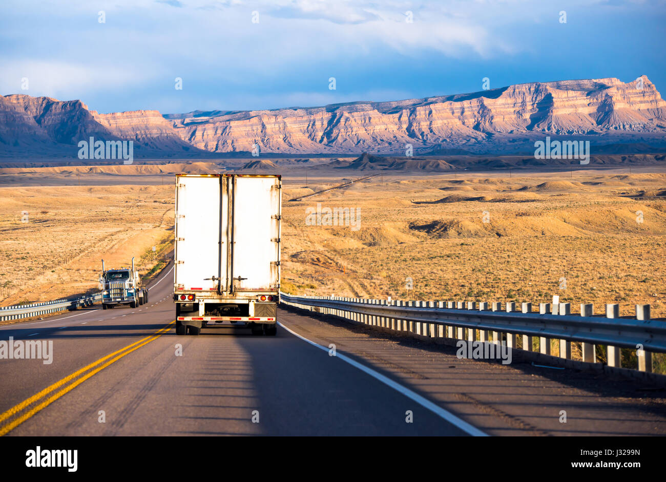 Semirimorchi con furgone a secco e rimorchi a letto piano che trasportano il carico sull'autostrada con recinzioni di sicurezza nello Utah nelle catene montuose illuminate dal sole Foto Stock