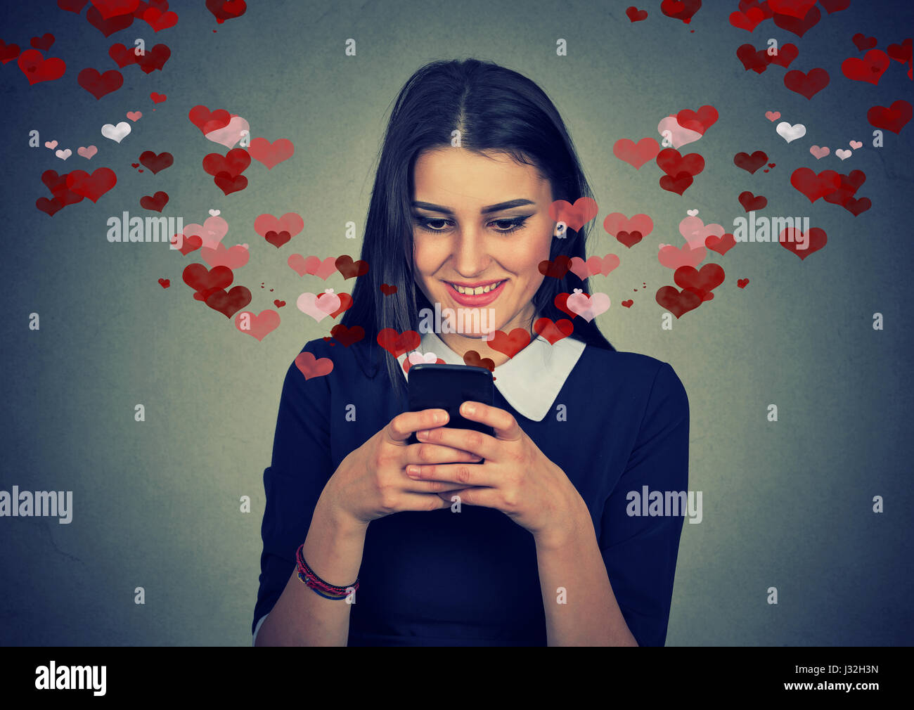 Ritratto di donna felice amore invio messaggio di testo sms sul cellulare con cuori rossi volare lontano dallo schermo isolato sul muro grigio Sfondo. Emo umana Foto Stock