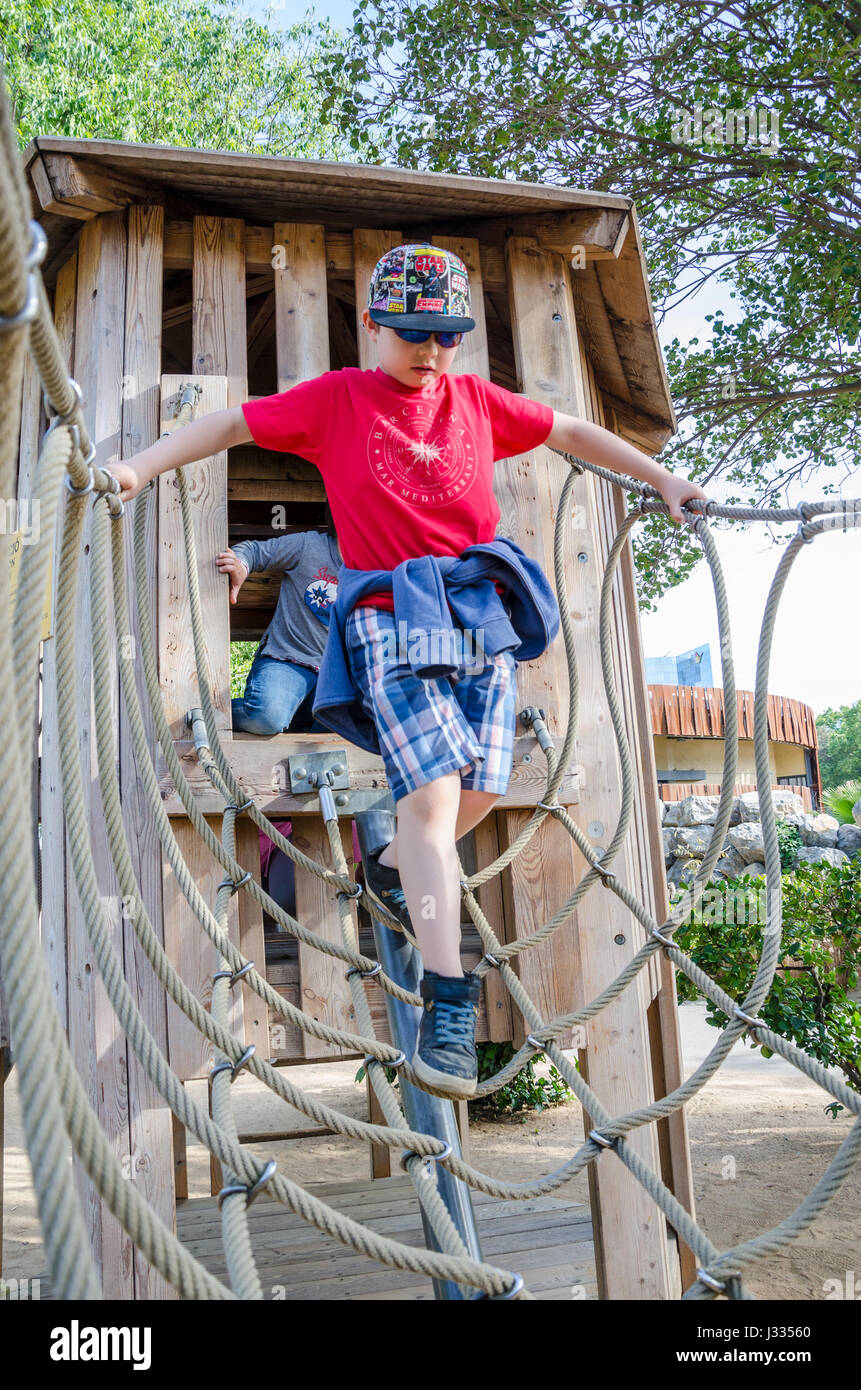 Un giovane ragazzo attraversa un ponte di corde che è parte di un telaio di arrampicata in un parco giochi per bambini. Foto Stock