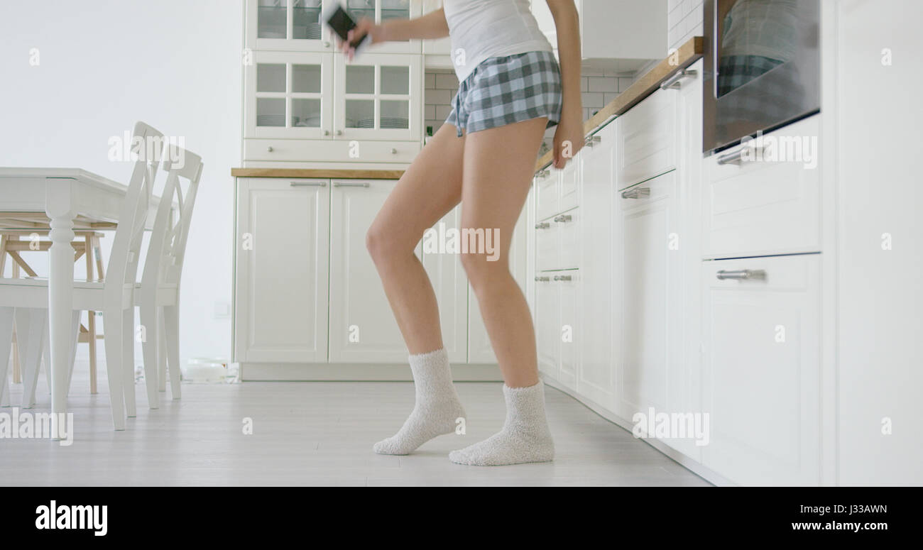 Raccolto di colpo senza volto di donna in home shorts tenendo il telefono in mano e danze nella cucina moderna. Foto Stock