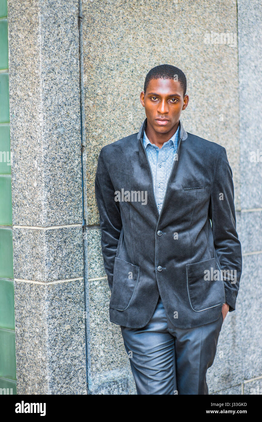 Giovane americano africano uomo che indossa di nuovo stile blazer nero, due mani mettendo in tasche, in piedi contro la parete sulla strada di New York, seriamente lookin Foto Stock