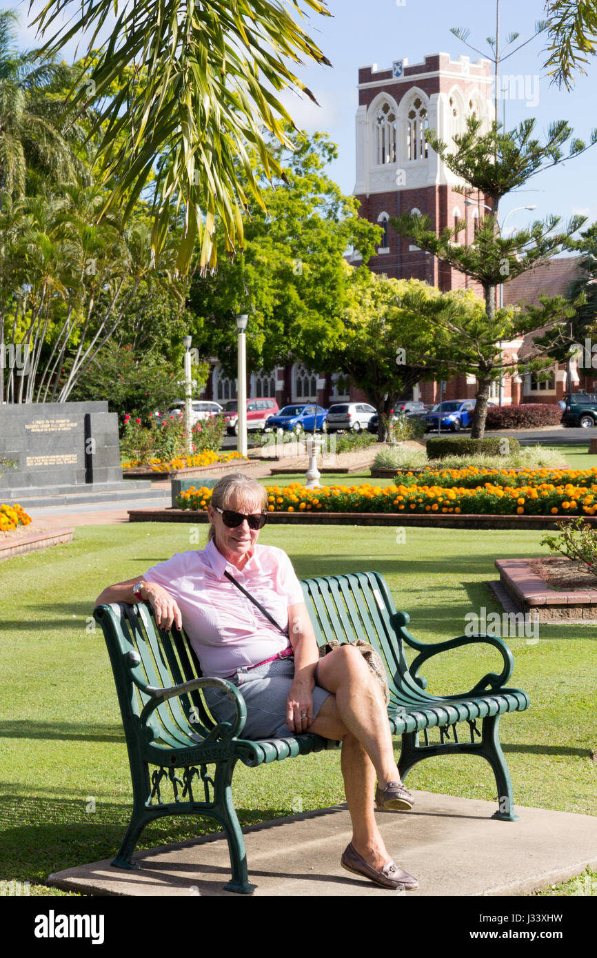 Senior donna sat in Buss park con la ex Presyterian St Andrews chiesa (ora la Chiesa Avventista del Settimo Giorno) in background, Bundaberg, Queenslan Foto Stock